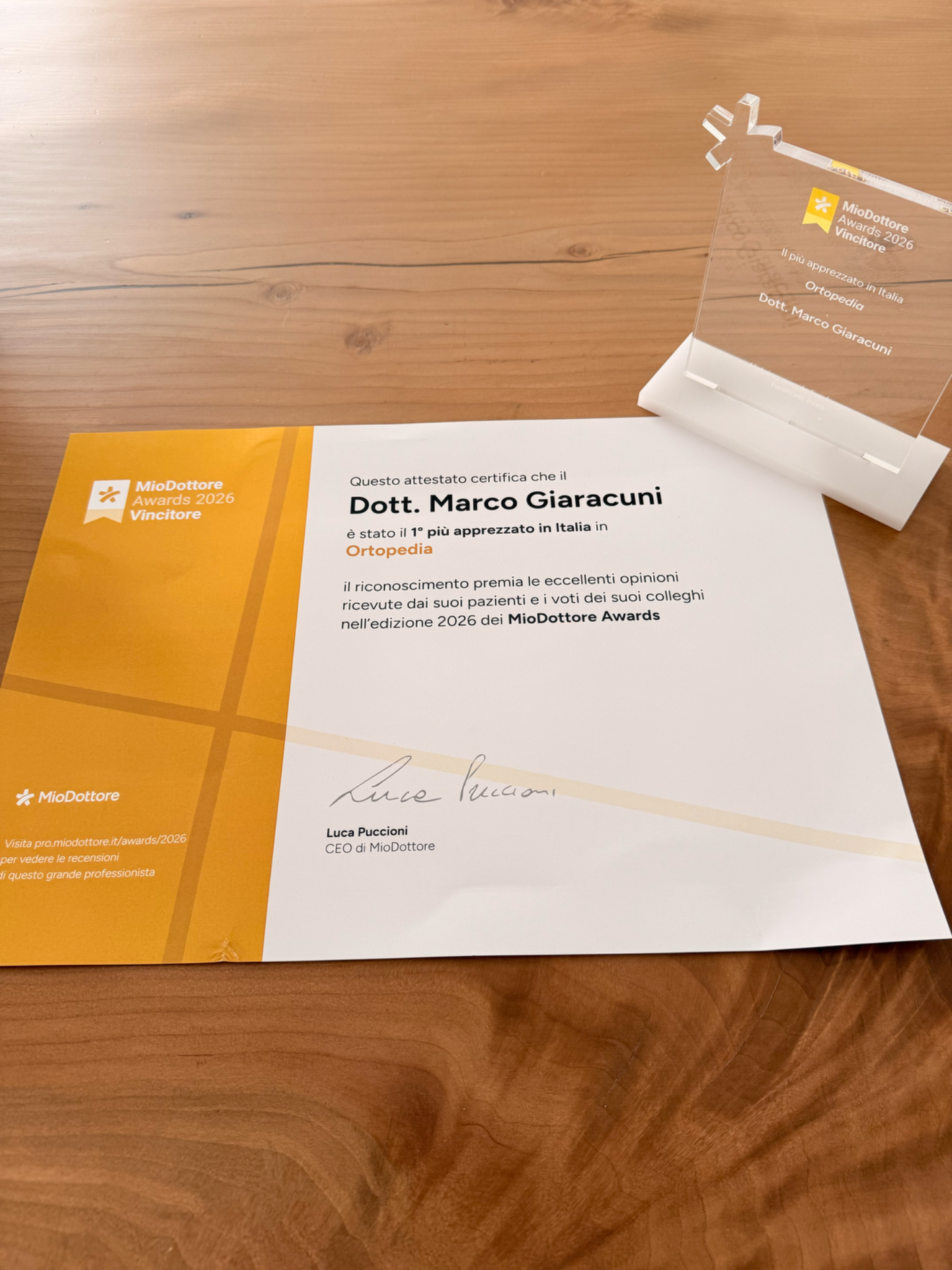 Attestato e brochure MioDottore Awards 2026 su tavolo di legno, con targa trasparente in alto.